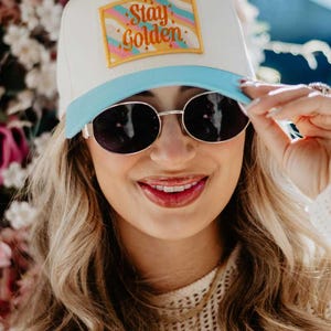 May include: A white baseball cap with a light blue brim and a rectangular patch that reads "Stay Golden" in gold lettering. The patch has a colorful, wavy design. The person in the image is wearing sunglasses.