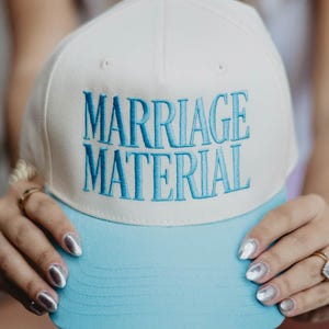 May include: A white and light blue baseball cap with the text "MARRIAGE MATERIAL" embroidered in blue. The cap has a curved brim and a button on top. A fun accessory for a wedding or engagement.