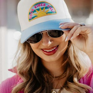 May include: A white and light blue baseball cap with the text "GOOD VIBES ONLY" in a colorful design. The cap has a curved brim and is worn by a person wearing sunglasses.