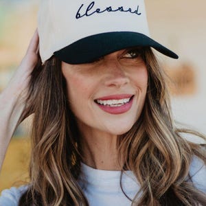 May include: Cream-colored baseball cap with a black brim. The word "blessed" is embroidered in navy blue script across the front. A casual, everyday accessory.
