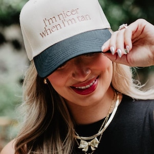 May include: A beige and navy baseball cap with the embroidered text "I'm nicer when I'm tan." The cap has a curved brim and a black button on top. The image shows a person wearing the cap.