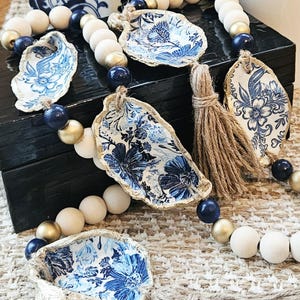 May include: Decorative beaded garland featuring oyster shells with blue and white floral patterns. The garland includes wooden beads in white, gold, and navy blue, and a jute tassel. The shells have a gold trim.