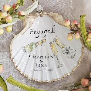 Op de afbeelding: Een wit sint-jakobsschelp ornament met goudkleurige randen, voorzien van een handgeschilderde illustratie van een champagnefles en twee glazen. De tekst luidt Engaged! CRISTIÁN & LIZA September 13, 2025. Het hangt aan een zilverkleurige kralenlus.