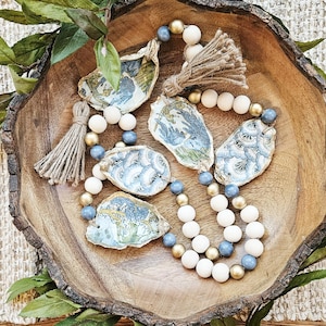 May include: A decorative garland featuring four oyster shells with blue and white floral patterns, interspersed with round wooden beads in matte white, metallic gold, and dusty blue. Two jute tassels are attached to the ends of the string.