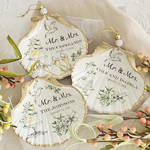 May include: Three decorative oyster shell ornaments with gold-painted edges and hanging loops. Each shell features a hand-painted wedding cake, a flying dove, floral accents, and personalized text including Mr. & Mrs., family names, and wedding dates.