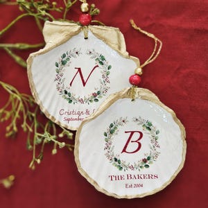 May include: Two white seashell ornaments with gold trim and red accents. Each ornament features a floral wreath design with a letter and text. One says "Cristian & September" and the other says "THE BAKERS Est 2004".