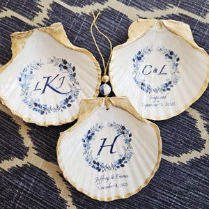 Pode incluir: Três conchas brancas em forma de pratos para anéis, com bordas douradas e desenhos florais azuis. Cada concha tem iniciais e datas, incluindo "C&L Engaged September 13, 20025", "L K J", e "H Jeffrey & Emma November 8, 2025". Uma corda dourada com contas conecta as conchas.