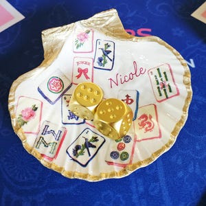 Può includere: Una conchiglia di capasanta bianca con bordi dipinti in oro, decorata con illustrazioni colorate di tessere del mahjong e il nome Nicole scritto in corsivo rosso. Due dadi dorati opachi sono posizionati al centro della conchiglia.