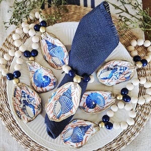 Könnte beinhalten: Ein weißer Teller hält eine marineblaue Serviette und muschelförmige Serviettenringe. Die Ringe weisen blaue Muscheldesign mit goldenen Verzierungen auf und sind mit weißen und marineblauen Perlen akzentuiert. Das Gedeck befindet sich auf einem gewebten Platzset.