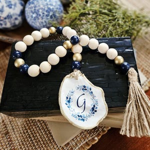 May include: A decorative garland featuring natural wood, navy blue, and metallic gold beads strung together. An oyster shell hangs from the strand, painted with a blue floral wreath surrounding the letter G, finished with a jute fiber tassel.