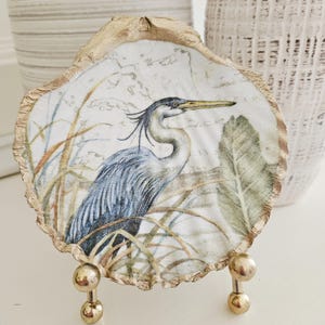 May include: A decorative oyster shell featuring a detailed illustration of a blue heron standing among tall grasses and large leaves. The shell has a scalloped edge painted in metallic gold and is displayed on a small gold metal stand.