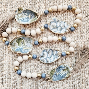 May include: Decorative garland featuring oyster shells with blue and white patterned designs, interspersed with wooden beads in natural, gold, and blue hues. The garland is finished with jute tassels.