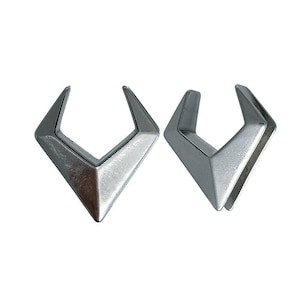 May include: A pair of silver-toned metal ear gauges featuring a sharp, angular V-shape design. Each piece has a smooth, polished surface with a pointed bottom and two upward-extending arms, creating a distinct geometric silhouette for ear stretching.