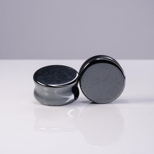 May include: Two circular ear plugs with a smooth, polished dark gray metallic finish. The plugs feature a concave side profile and flat circular faces, reflecting light on their glossy surfaces. These body jewelry pieces are solid and opaque.