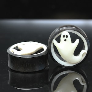 May include: Pair of black wood ear gauges with a white ghost design carved into the wood. The gauges are round and have a smooth finish.