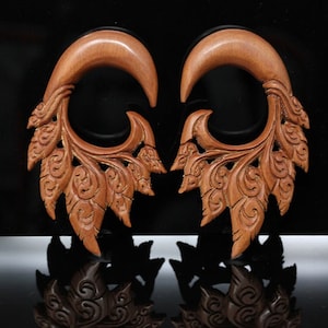 May include: A pair of wooden ear gauges with intricate carvings in the shape of leaves and swirls. The gauges are a rich brown color and have a polished finish.