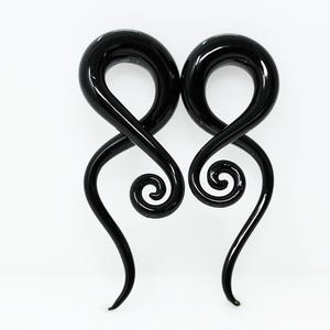 May include: A pair of black ear tapers featuring a smooth, glossy finish. Each piece has a circular top section that transitions into a tapering, elongated spiral shape that curves downward, creating a continuous, fluid, and symmetrical design.