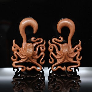 May include: A pair of wooden octopus ear gauges. The gauges are carved in the shape of an octopus with detailed tentacles. The gauges are a reddish-brown color.