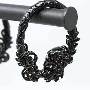 May include: A pair of black hoop earrings featuring an intricate design of a skull surrounded by sculpted roses and swirling vine patterns. The earrings are crafted with a polished finish and are displayed hanging from a horizontal black bar.