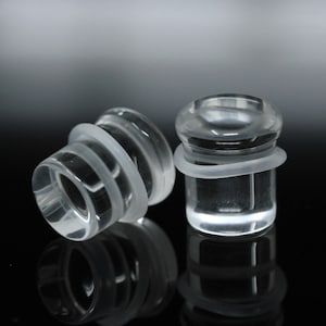May include: Two clear glass ear gauges with a white rubber o-ring on each. The gauges are cylindrical and have a smooth, polished finish.