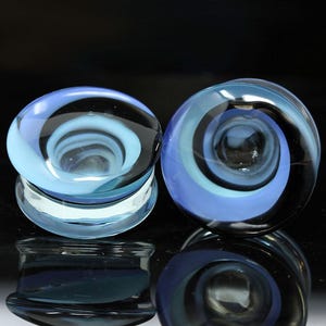 May include: A pair of circular glass ear plugs featuring a swirling pattern of translucent light blue, dark blue, and black colors. The plugs have a smooth, polished surface with a concave center and a flared edge design for secure placement.