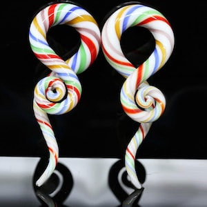 May include: A pair of glass ear tapers featuring a swirling candy cane pattern with white, red, yellow, green, and blue stripes. Each piece has a circular loop at the top that transitions into a tapered, curved tail, creating a spiral shape.