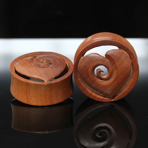 May include: Two wooden ear gauges with a heart-shaped design carved into the wood. The heart has a swirl pattern inside.