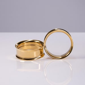 May include: Two gold-colored ear tunnels with a smooth, polished finish. One tunnel is positioned horizontally, showing its cylindrical shape and flared edges, while the other stands vertically, displaying its circular opening and reflective surface.
