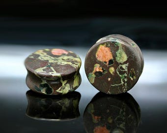 May include: A pair of circular ear plugs crafted from a dark brown stone featuring natural inclusions of mottled green, black, and salmon pink. The plugs have a smooth, polished finish with a concave profile, showing distinct mineral patterns.