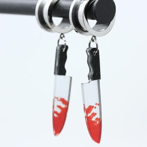 May include: A pair of silver-toned circular ear tunnels featuring dangling knife-shaped charms. Each charm has a black handle and a clear blade section with a red splatter pattern resembling blood, suspended from the tunnels by small metal jump rings.