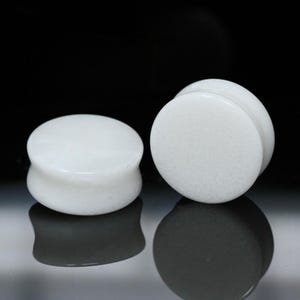 May include: A pair of solid white circular ear plugs with a smooth, polished finish. The plugs feature a classic saddle-style design with flared edges, creating a clean and uniform appearance. These circular body jewelry pieces are opaque white.