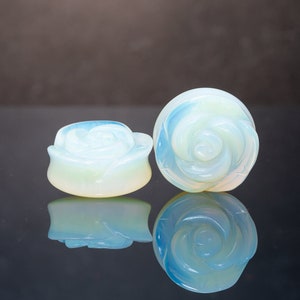 May include: Two opalite rose plugs, one larger than the other, are shown on a black surface. The plugs are a light blue color with a white center.