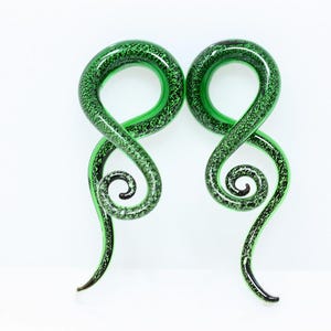 May include: A pair of spiral-shaped ear gauges made of translucent green glass with a speckled black pattern throughout. Each piece features a circular top section that tapers into a curved, pointed tail, creating a distinct swirling silhouette.