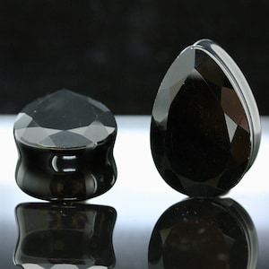 May include: Two black, faceted plugs for stretched ear piercings. One is a rounded shape, the other is teardrop-shaped. Both have a glossy, reflective surface, and are displayed against a dark background.