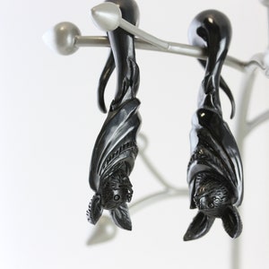May include: Two black bat-shaped earrings hanging from a silver stand. The earrings are made of wood and have intricate details.