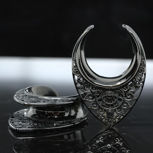 May include: Two black metal ear gauges with intricate, ornate designs. The gauges are shaped like a crescent moon and a half-circle.