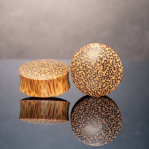 May include: Two wooden ear gauges with a brown and black speckled pattern. The gauges are round and have a slightly flared shape.