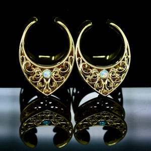 May include: A pair of gold-toned ear weights featuring a crescent-shaped design with intricate filigree metalwork and a single round, iridescent opal-like stone centered at the base of each piece, reflecting light against a dark surface.