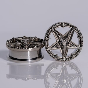 May include: A pair of silver-toned metal ear plugs featuring a circular design with a central pentagram and a goat skull motif. The outer ring is decorated with small skull accents, and the cylindrical body has a smooth, polished finish.