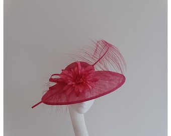 May include: A bright pink wide-brimmed hat with a large pink flower and a feather accent. The hat is on a white mannequin head. The text "Jayne Alison Millinery" is in the lower left corner of the image.