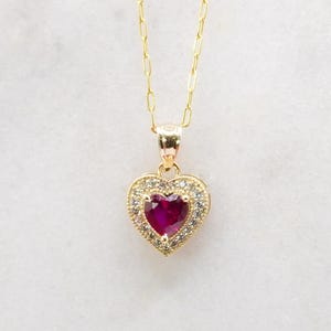 May include: A gold necklace with a heart-shaped pendant. The pendant features a ruby-colored center stone surrounded by a halo of smaller, clear stones. The chain is delicate. The text "POSH GOLD EST.2013" is at the bottom.