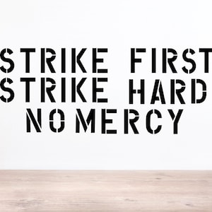 May include: A black wall decal featuring the text STRIKE FIRST, STRIKE HARD, NO MERCY in a bold, stencil-style font. The lettering is arranged in three distinct lines, creating a sharp contrast against the plain white background.