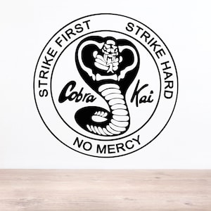 May include: A circular black and white decal featuring a central cobra snake illustration. The text "STRIKE FIRST", "STRIKE HARD", and "NO MERCY" is printed along the circular border, with the words "Cobra Kai" positioned across the center.