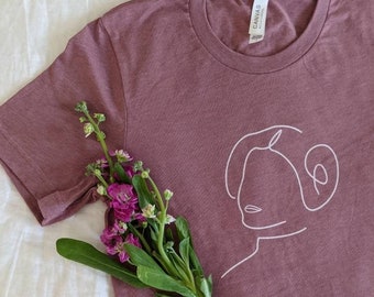 May include: A mauve t-shirt with a white outline of Princess Leia's face. The shirt is laying on a white surface with a bouquet of purple flowers.