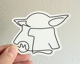May include: A white sticker with a black line drawing of a character from the Star Wars franchise, known as "The Child" or "Baby Yoda". The character is depicted in a standing pose, wearing a robe and holding a small object in its hand.