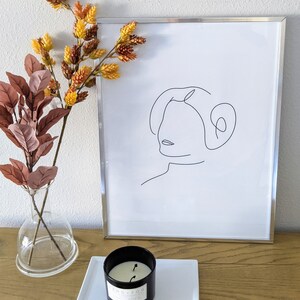 Pu&ograve; includere: Una stampa incorniciata in nero con un disegno a linea di un viso femminile. La stampa &egrave; su un tavolo di legno con un vaso di vetro trasparente con fiori secchi e una candela nera su un piatto quadrato bianco.