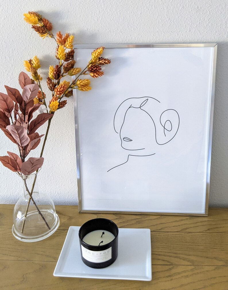 Pu&ograve; includere: Una stampa incorniciata in nero con un disegno a linea di un viso femminile. La stampa &egrave; su un tavolo di legno con un vaso di vetro trasparente con fiori secchi e una candela nera su un piatto quadrato bianco.