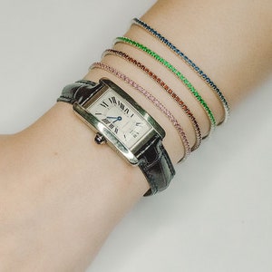 May include: A silver watch with a black leather band and a rectangular face. The watch is worn on a wrist with several colorful bracelets stacked on top of each other.