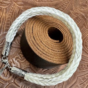 May include: A white braided bracelet with silver-toned metal clasps. The bracelet is made of a thick, woven material and is displayed on a brown leather surface. The clasp has an ornate design. A rolled-up piece of brown leather is in the background.