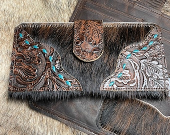 Könnte beinhalten: Eine braune Lederbörse mit einem Haarfell-Design und türkisfarbenen Akzenten. Die Börse hat einen Klappverschluss und ein dekoratives, geprägtes Blumenmuster.